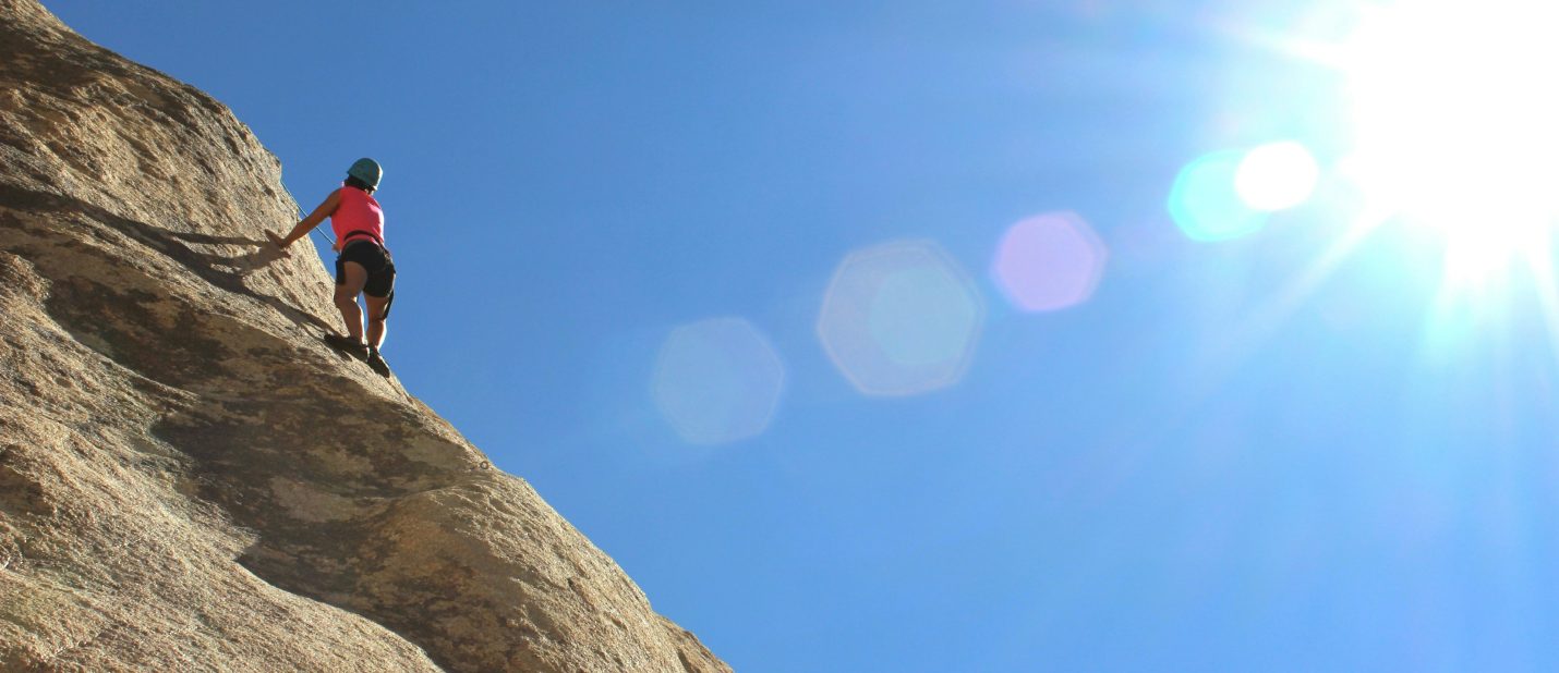 A person climbing up a rock face with a clear sky and bright sun in the background